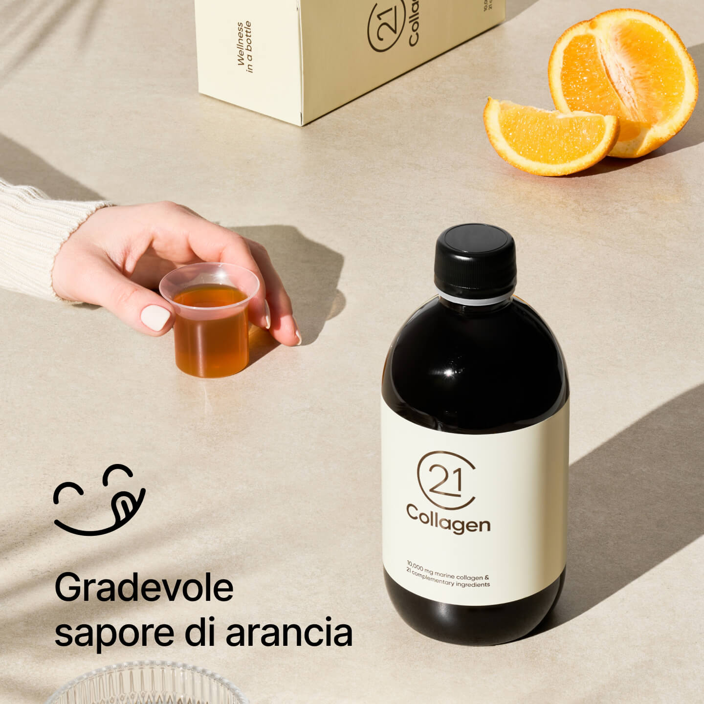 Mano con dose giornaliera di 21Collagen al gusto arancia; bottiglia e scatola sullo sfondo con fette d’arancia. 